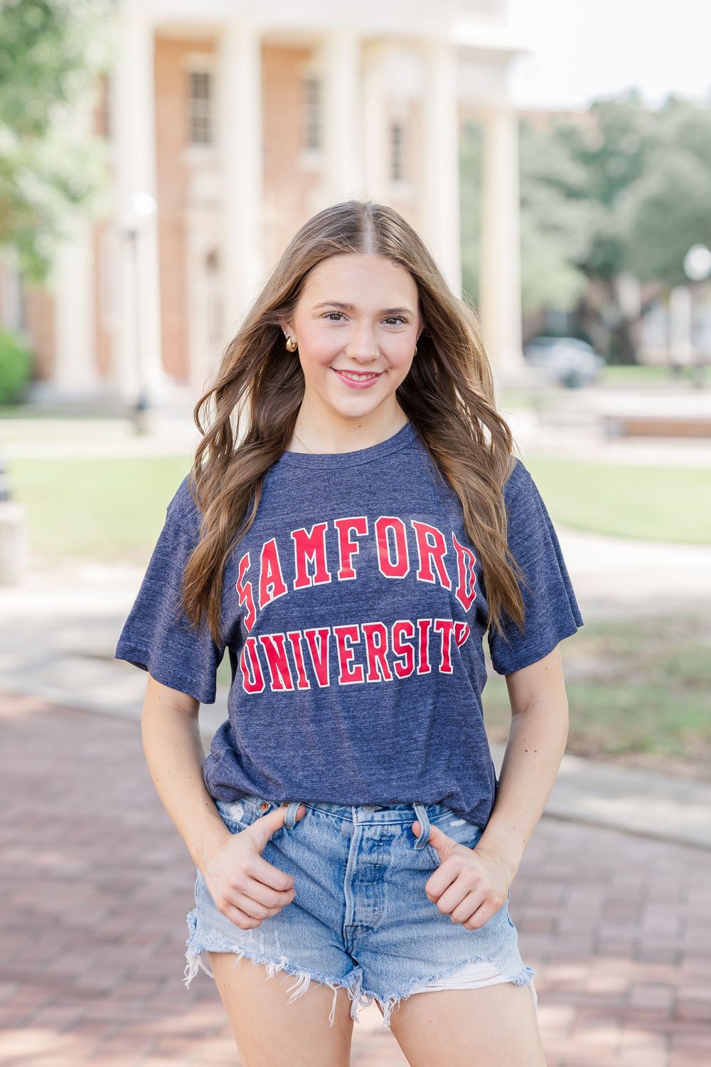 Person wearing a navy blue college t-shirt and distressed denim shorts poses outdoors on a brick walkway.
