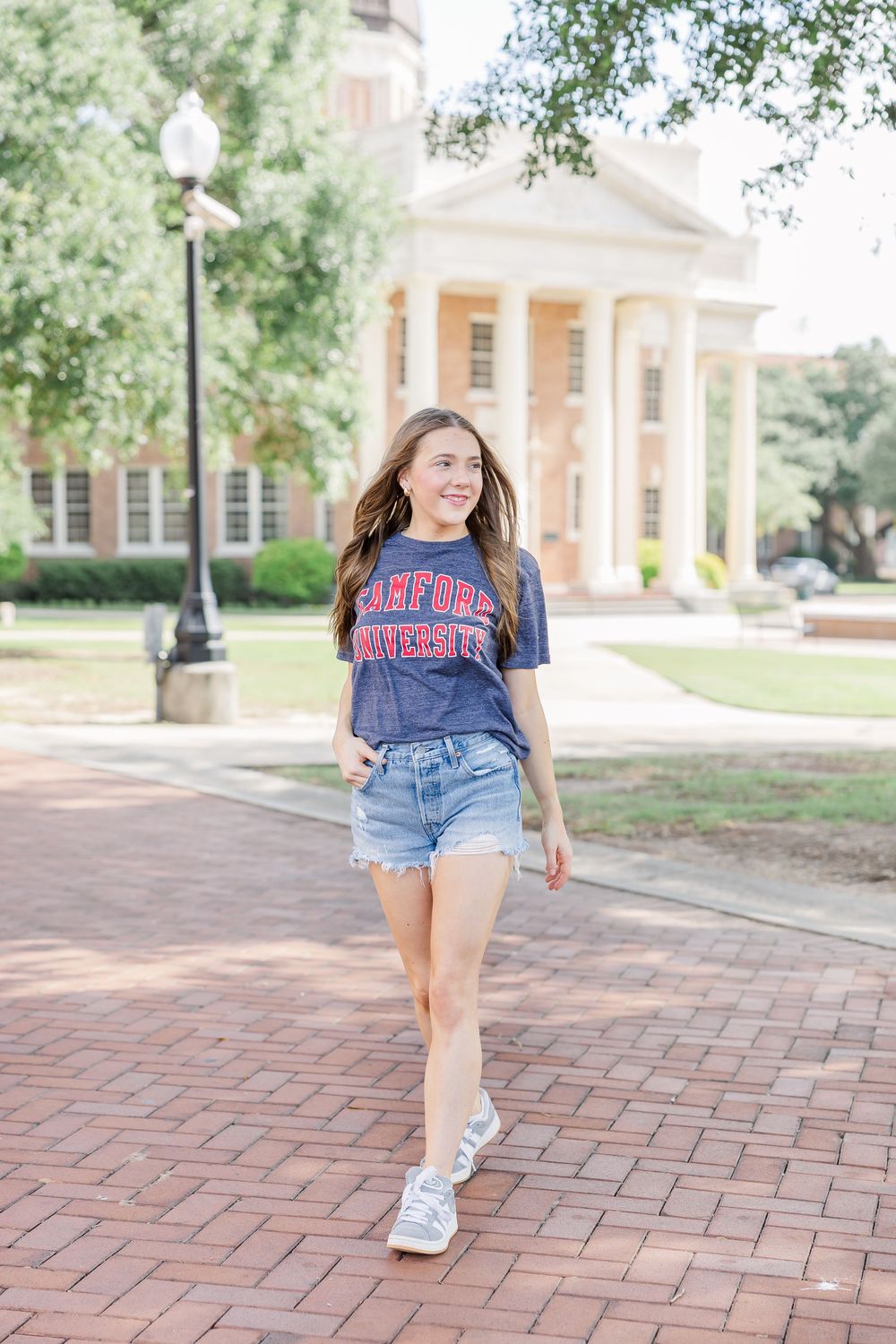 Student walking on brick path wearing Ole Miss university t-shirt and denim shorts near classic college building.
