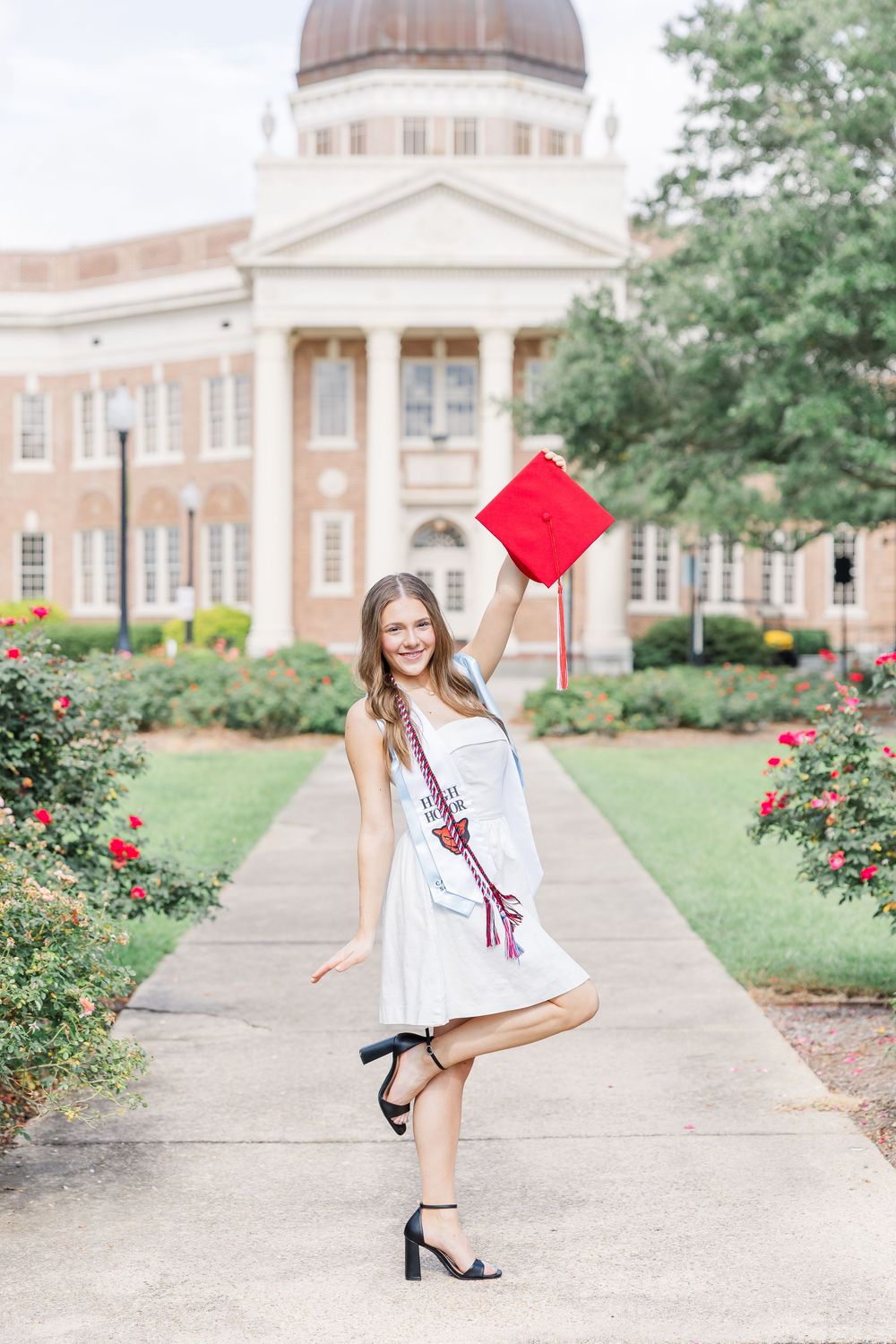 Graduate in white dress and red graduation cap poses playfully on campus walkway with roses and buildings in background.
