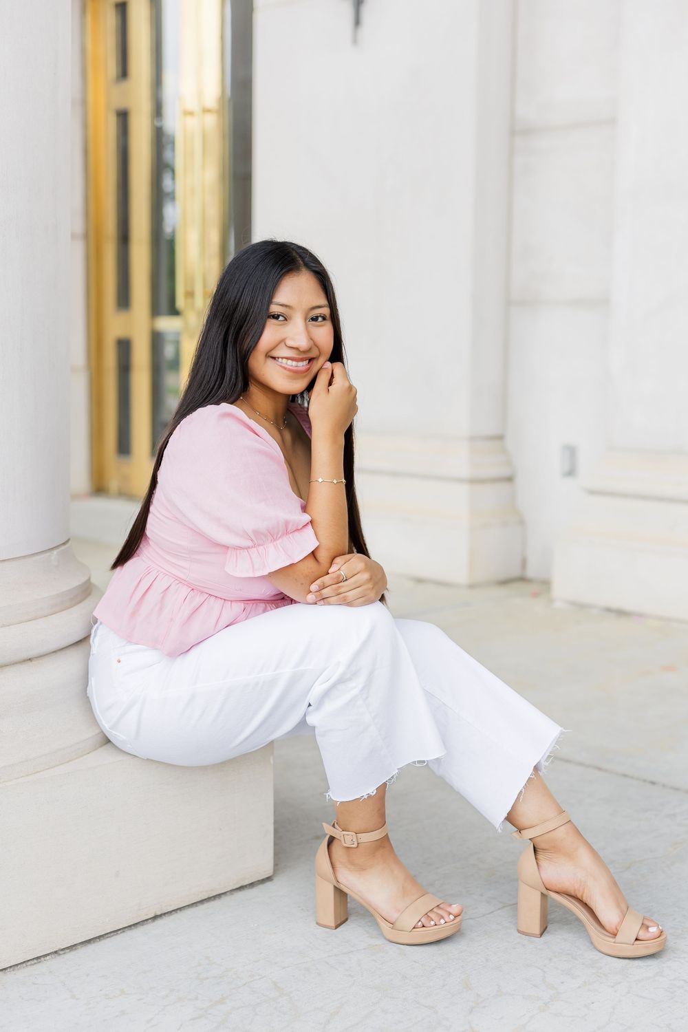 A person in a pink sweater and white pants sits elegantly on marble steps while wearing beige heeled sandals.