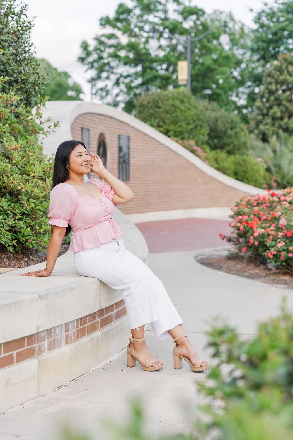 A person in a pink top and white pants sits gracefully on a concrete wall surrounded by blooming roses and greenery.