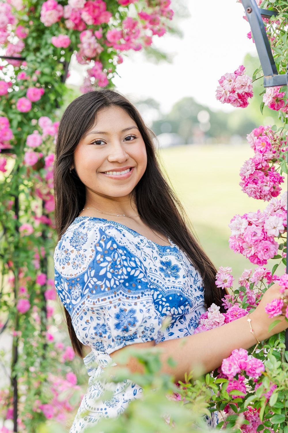 Young person in blue and white floral dress poses among vibrant pink flowering branches in outdoor spring setting.