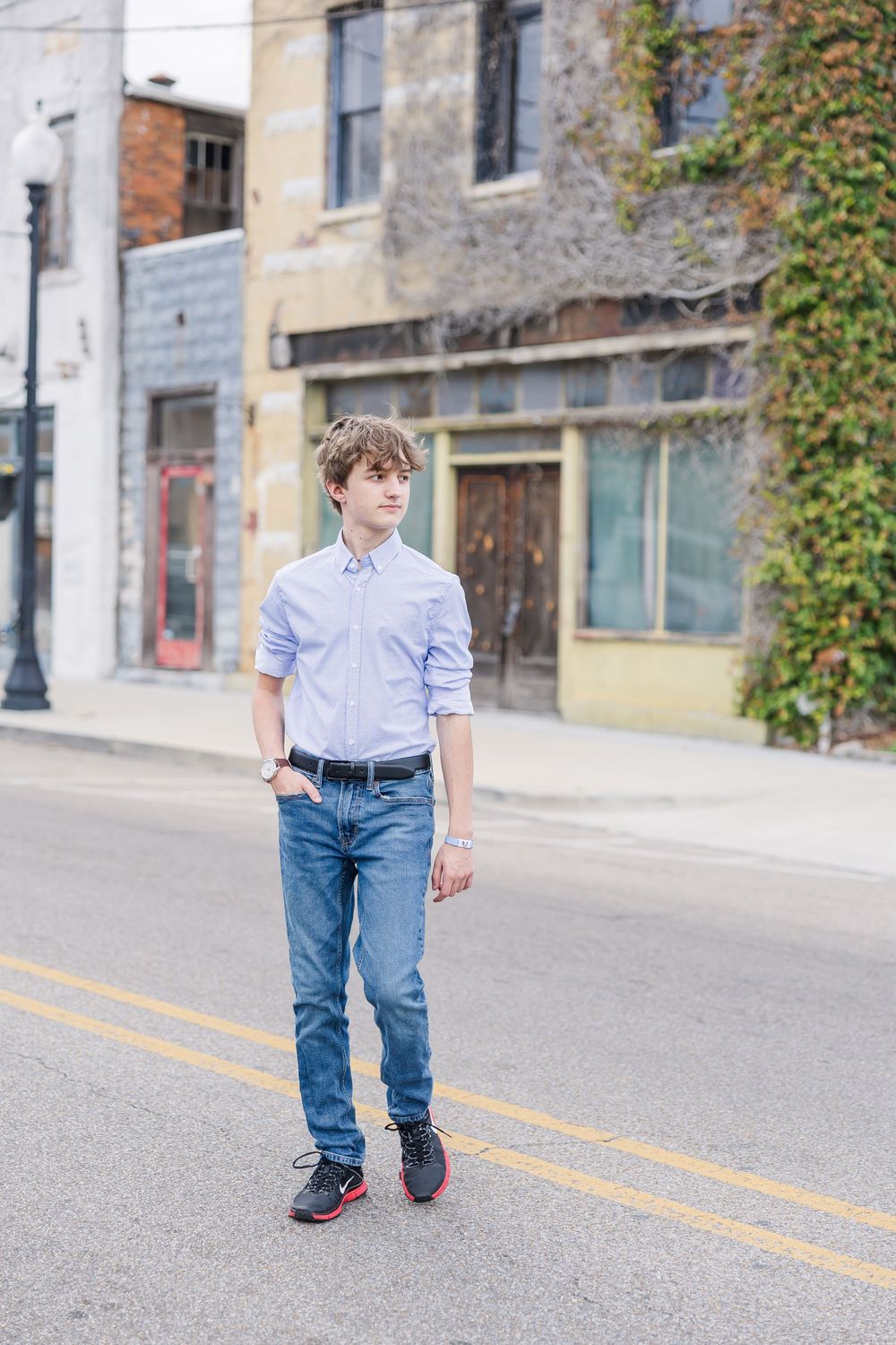 Someone in casual attire with a light blue button-down shirt and jeans stands on a street with old buildings in the background.