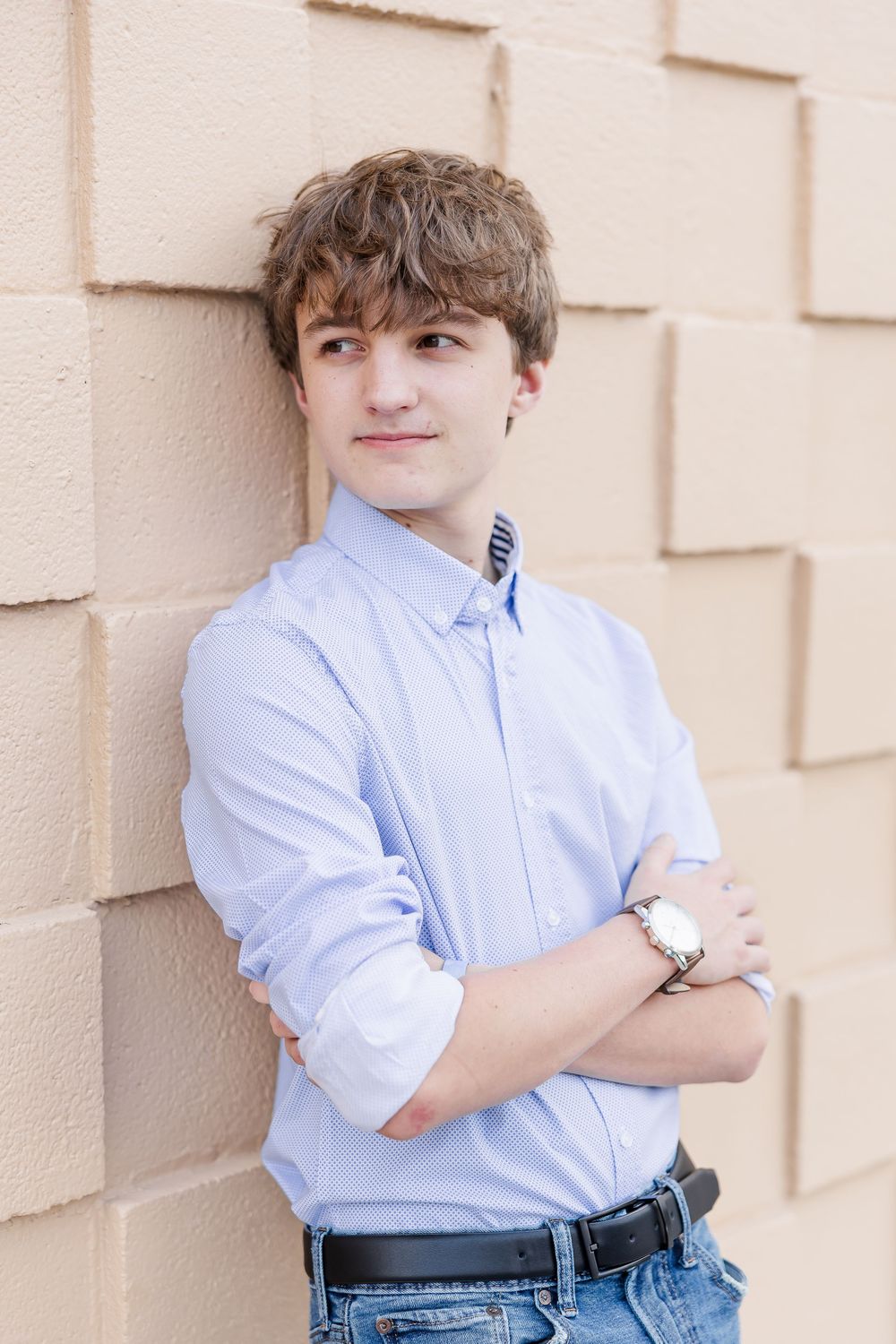 A young person in a light blue button-down shirt and jeans poses confidently against a beige textured wall.