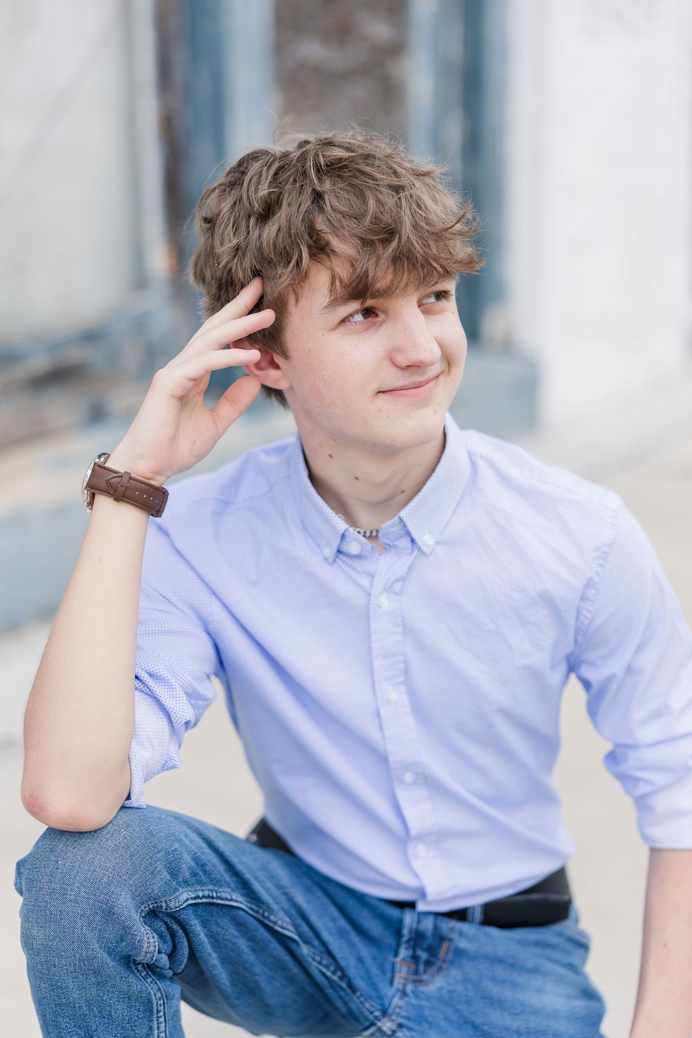 Young person in light blue button-down shirt and jeans poses casually outdoors with hand near ear.