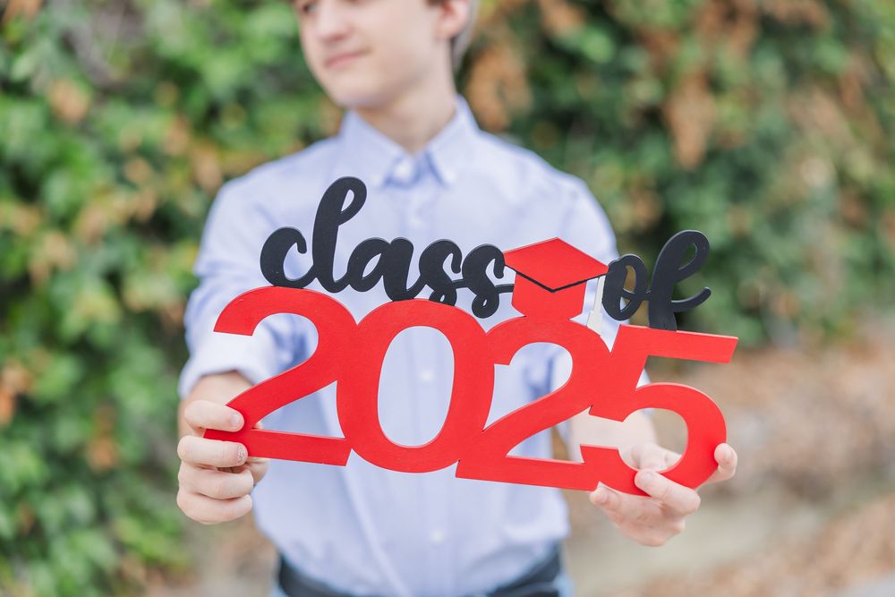 A person in a white button-down shirt holds red graduation numbers and letters spelling out 'Class of 2021' outdoors.