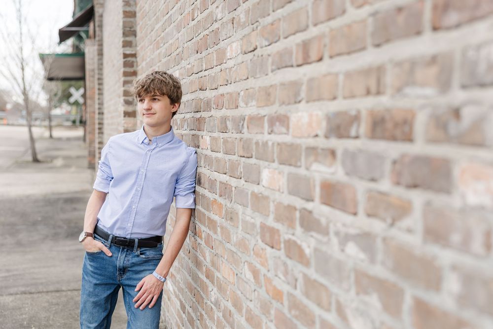 Person in light blue shirt and jeans poses casually against rustic brick wall in outdoor setting.