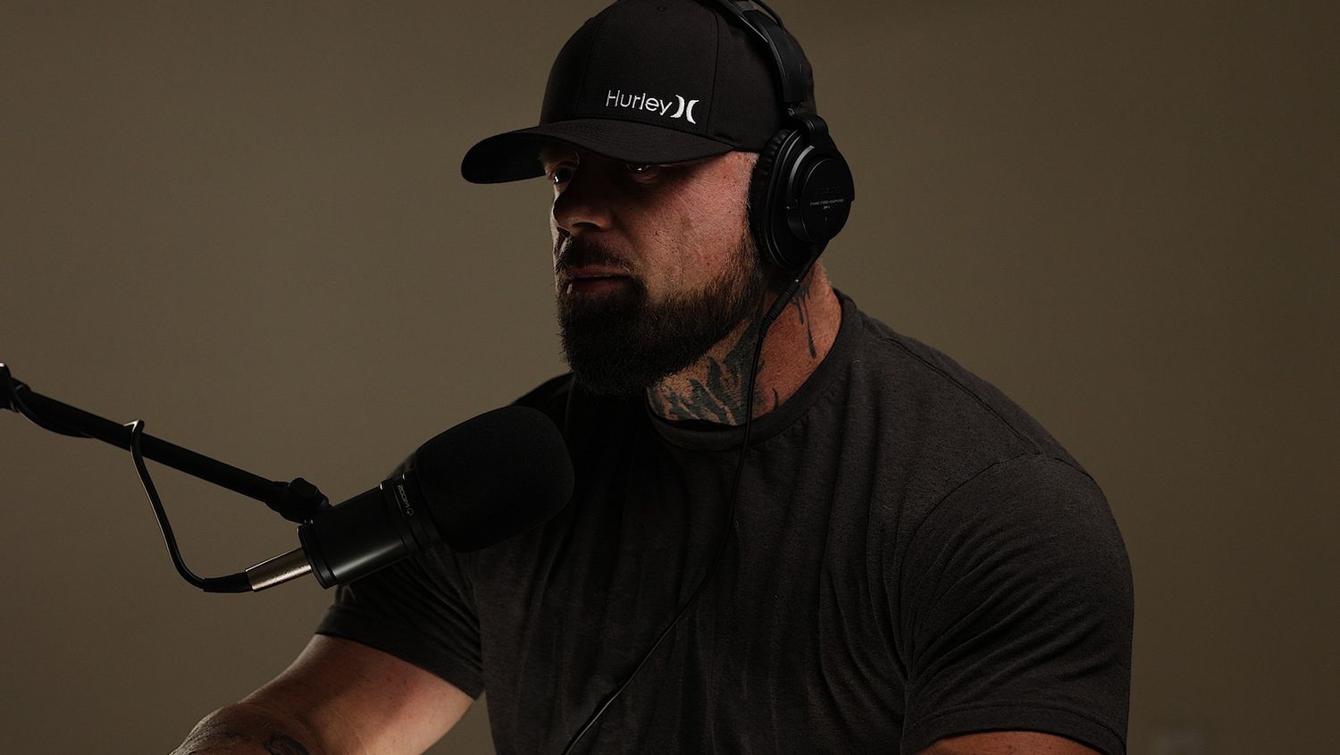 Someone in a black cap and headphones speaking into a podcast microphone in a dark studio setting.