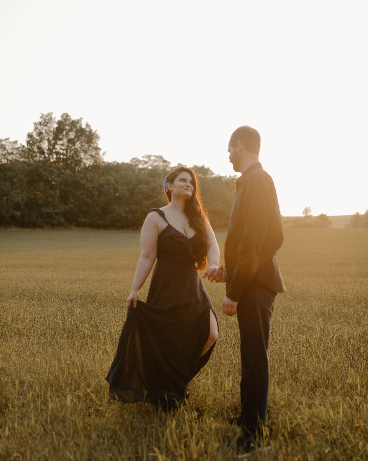 A couple stands together in a grassy field at sunset, with the person in a long black dress and their partner in dark attire.