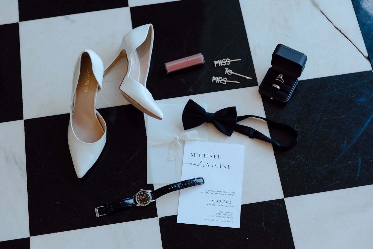 Wedding accessories including white heels, black bow tie, and rings on a checkered surface.