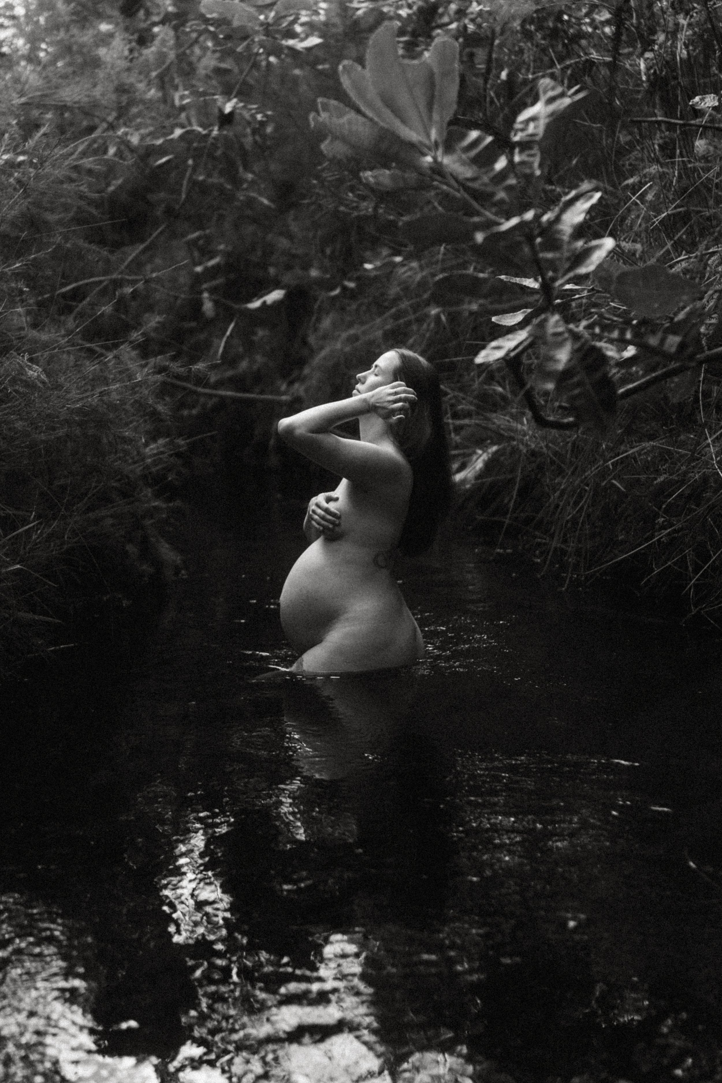 Artistic black and white photograph shows a pregnant woman, tranquil forest waters surrounded by dense foliage.