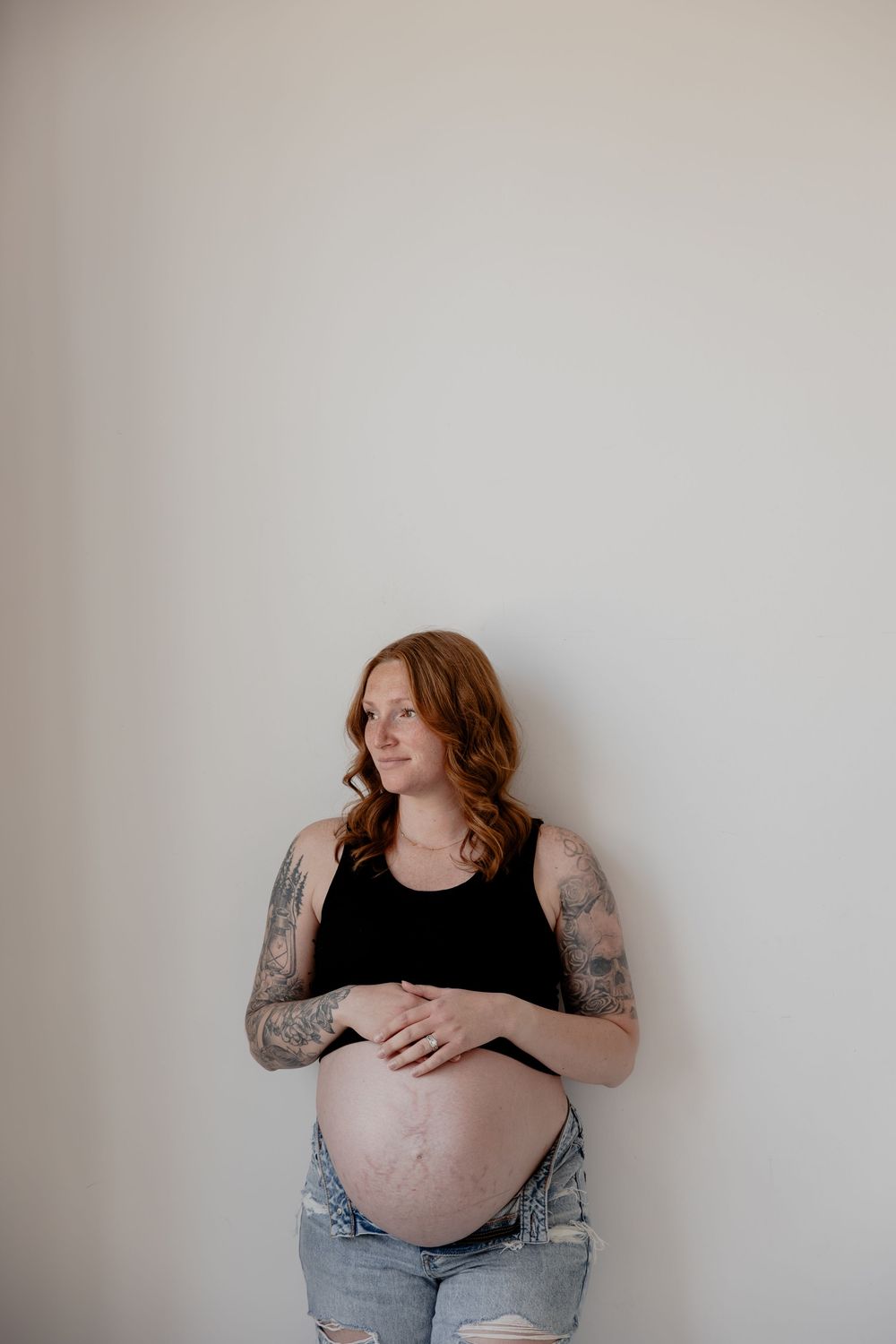 Maternity photoshoot featuring a woman with tattoos in a black tank top against white wall in Milwaukee, WI  with Essence Rose Photography.