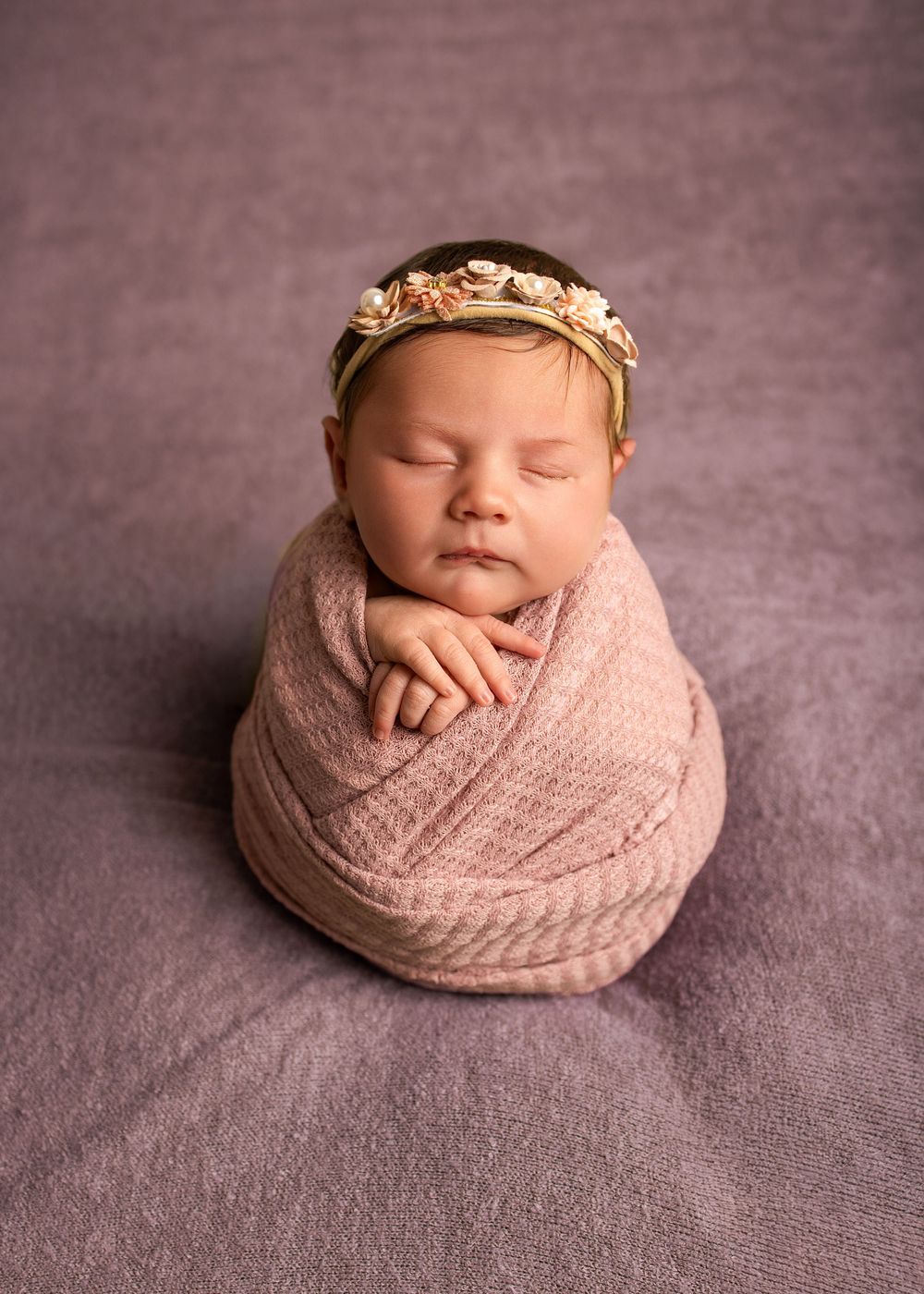 A sleeping newborn baby swaddled in soft pink knit fabric with a delicate gold leaf crown rests peacefully on a gray backdrop.