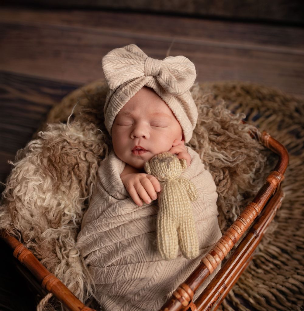 A sleeping newborn baby wearing a knit outfit lies in a rustic wooden basket with fluffy cream-colored bedding.
