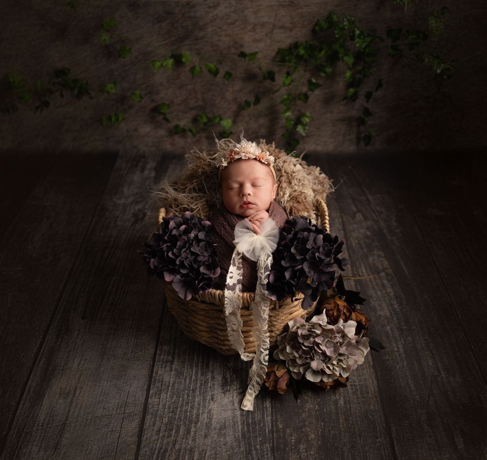 A sleeping baby in a rustic basket surrounded by dried flowers and wearing a delicate crown on a wooden floor.