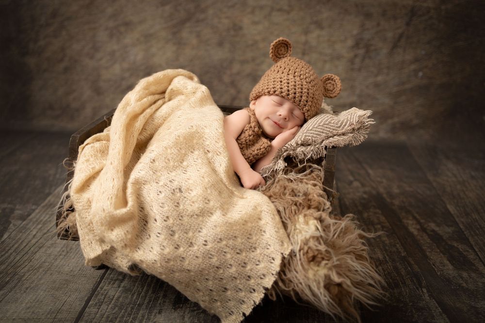 Newborn wearing brown bear hat sleeps peacefully on textured fur backdrop with cream lace blanket.