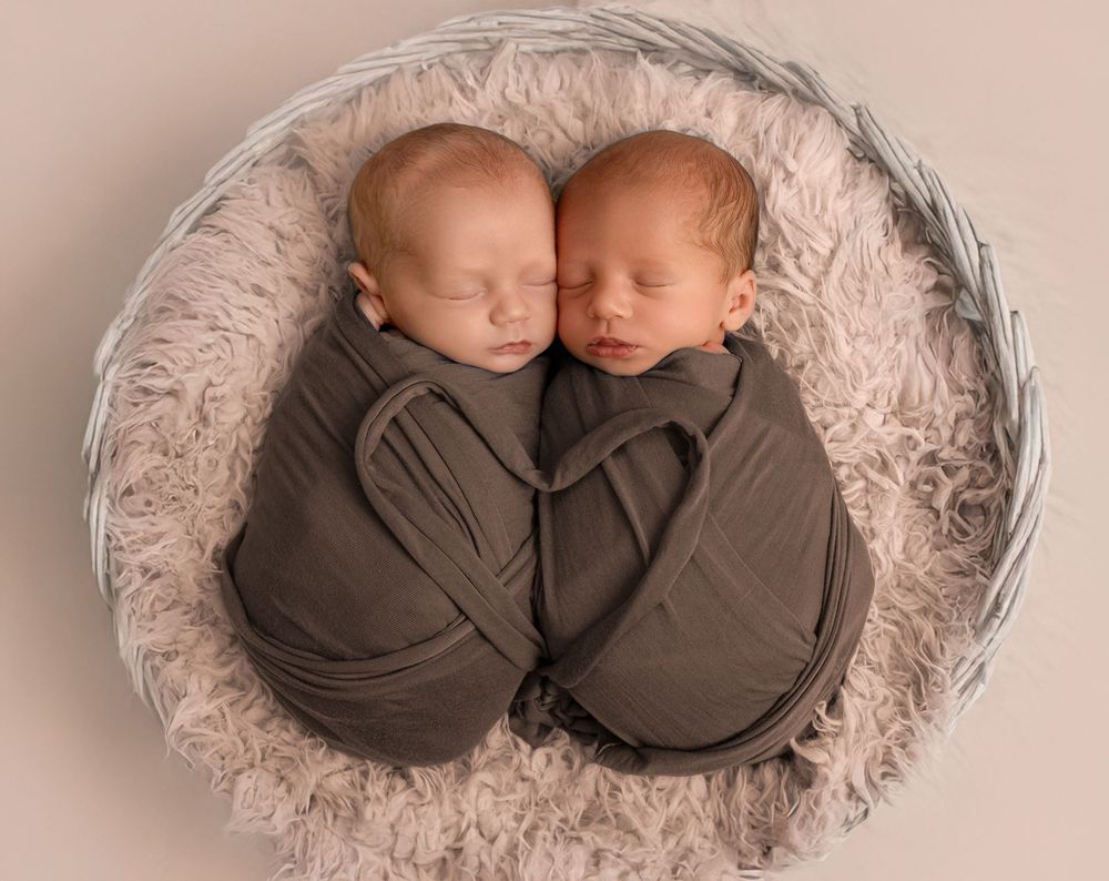 Two sleeping newborn babies in brown wraps lying together on soft beige fabric in various poses.