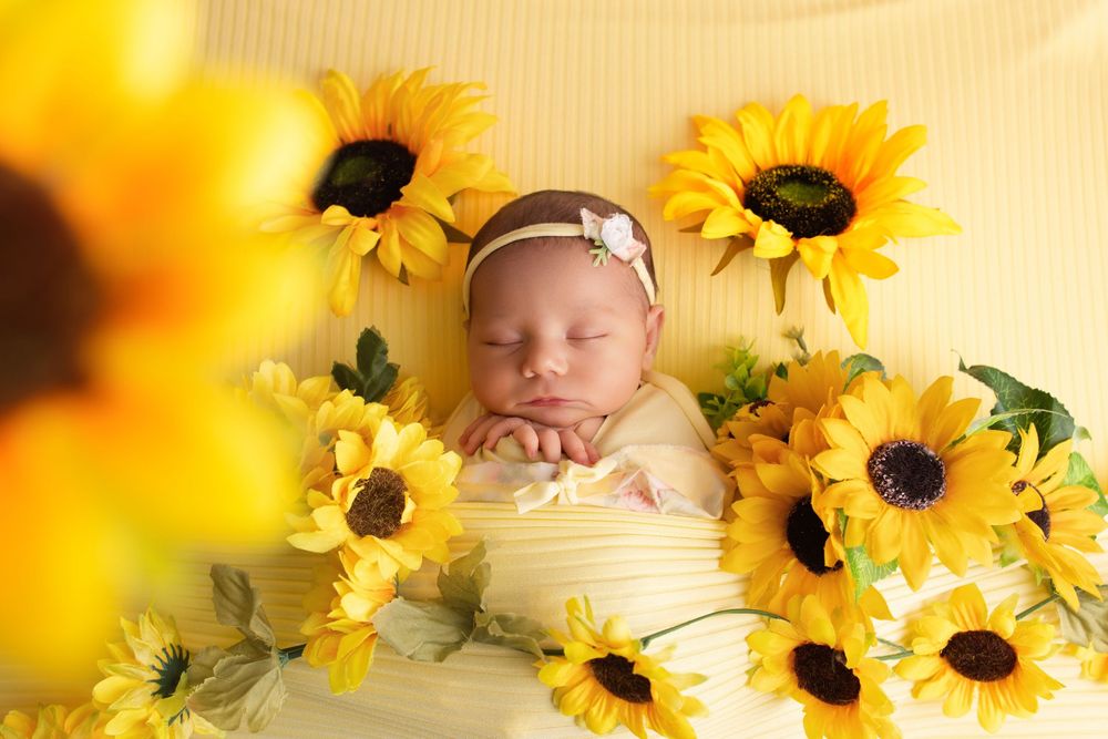 Sleeping newborn baby girl in yellow wrap surrounded by bright sunflowers, peaceful studio portrait.