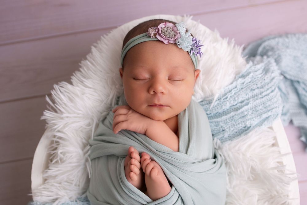 Sleeping newborn baby girl swaddled in teal wrap with floral headband resting in white basket with fluffy fur.