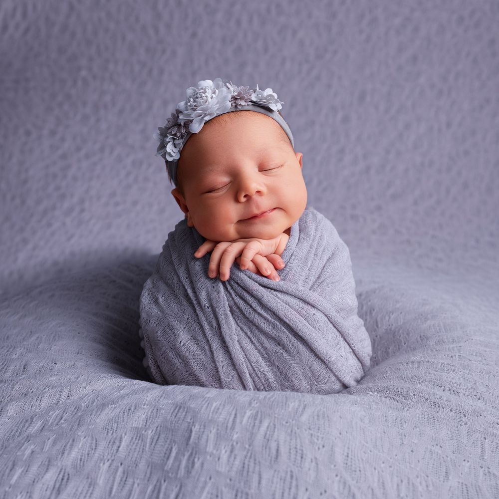 Smiling newborn baby swaddled in gray wrap with floral headband, posed on soft gray pillow.