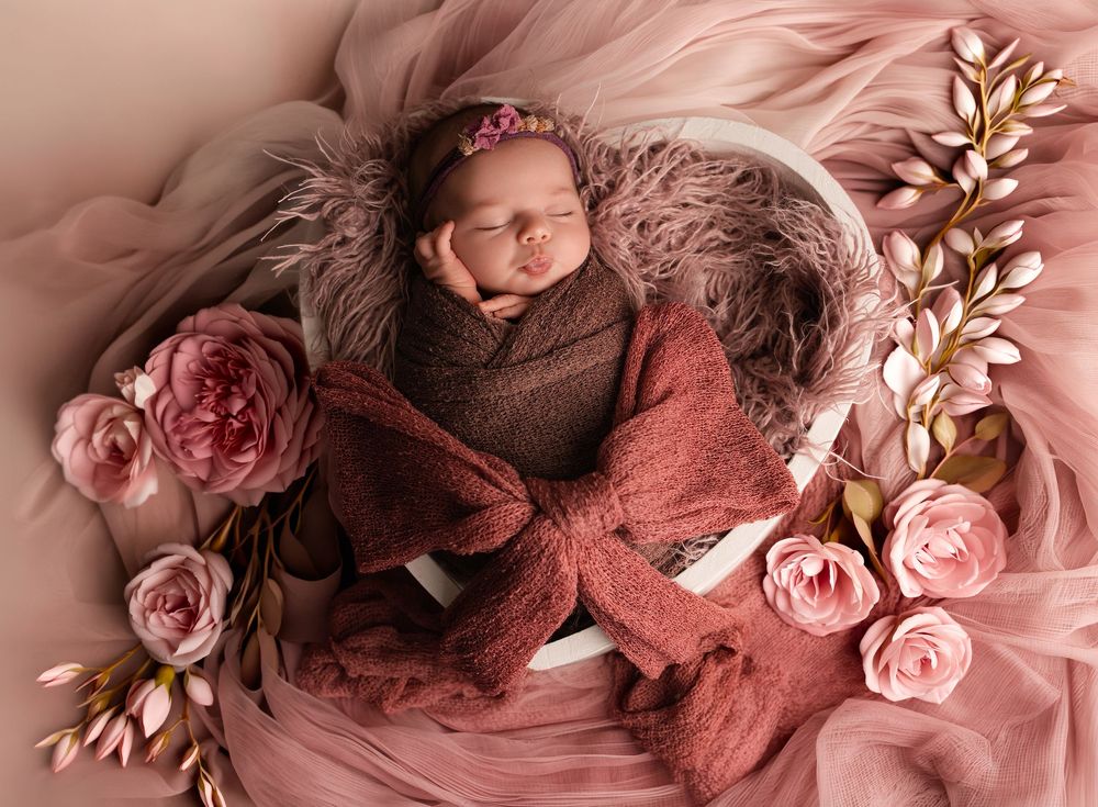 Newborn baby sleeping peacefully in a bowl surrounded by pink roses and soft mauve fur, wrapped in a dusty rose swaddle.