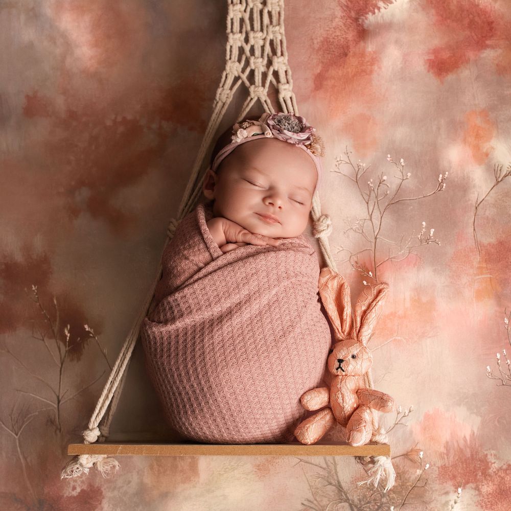 Sleeping newborn baby wrapped in pink knit, wearing floral headband, on macrame swing with bunny toy.