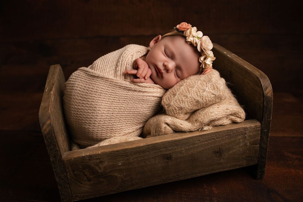Newborn baby swaddled in cream knit wrap with floral headband sleeping peacefully in a rustic wooden mini bed.