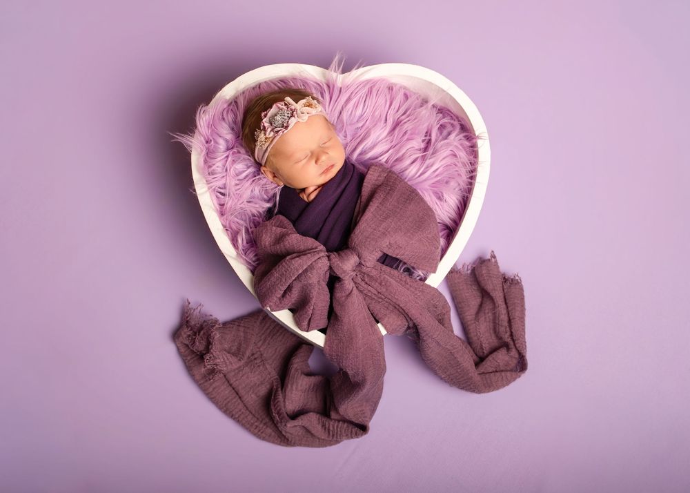 A collection of baby clothes and blankets arranged in a soft pink gradient against a purple background.