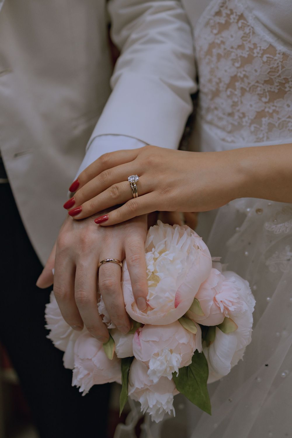 Close-up of hands with wedding rings resting on pink peonies bouquet.