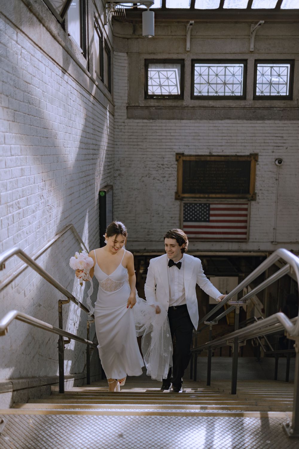 Two people in formal wear walk down stairs in a building with white brick walls and an American flag.