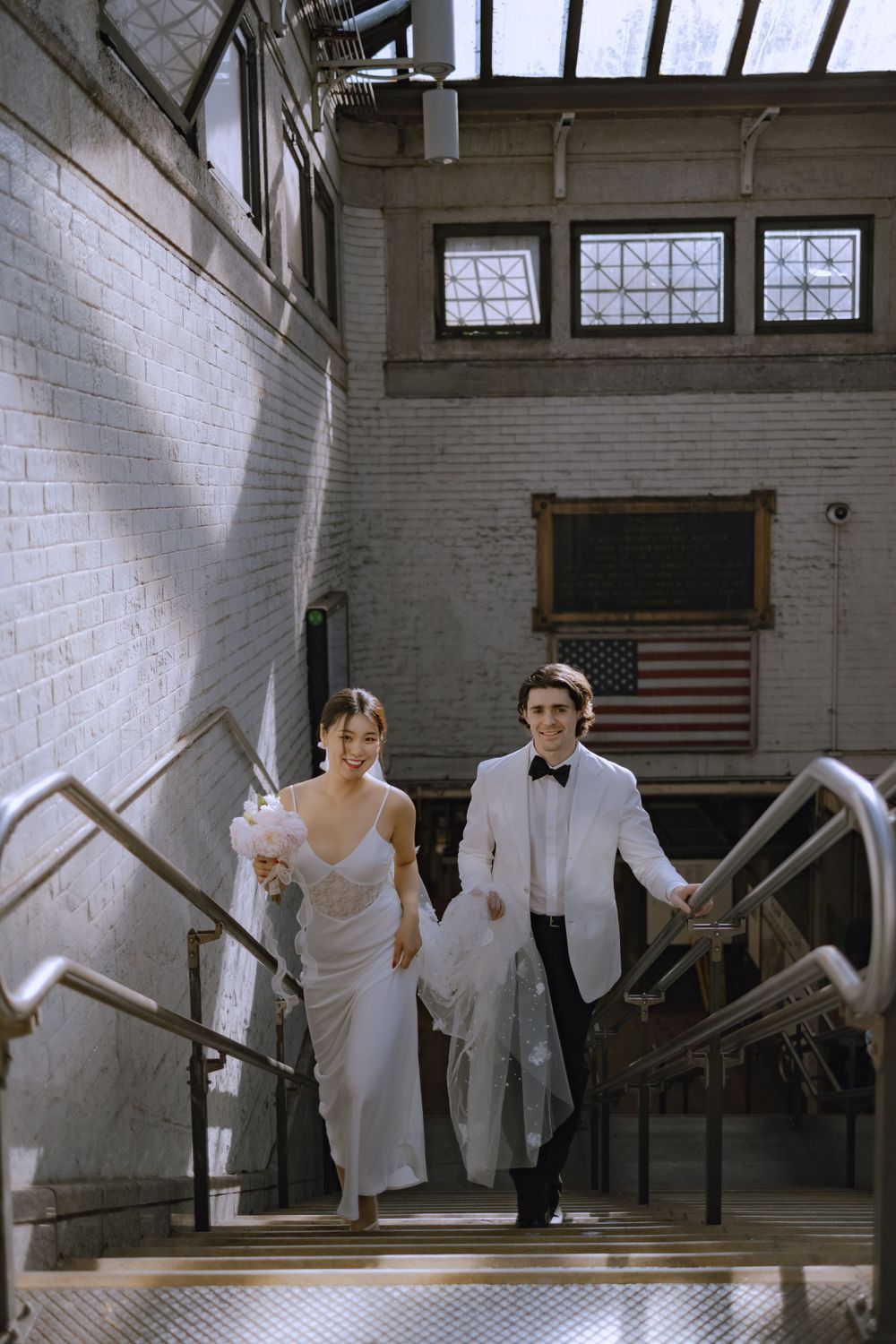 A couple in white formal wear descends concrete stairs in an industrial urban setting.