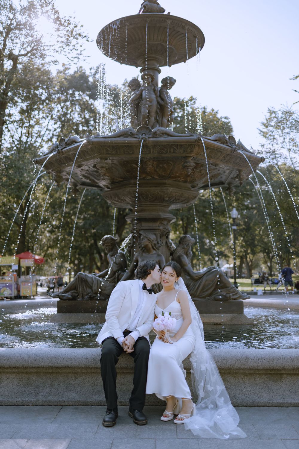 A couple in wedding attire sits and poses by an ornate fountain in a park.