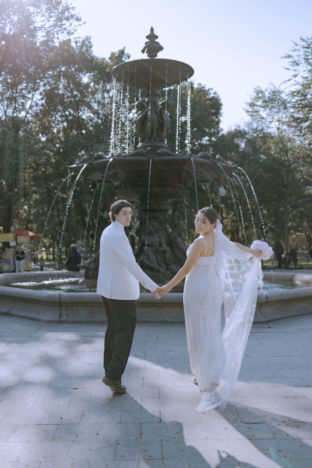 A couple in wedding attire dances playfully around a large fountain.