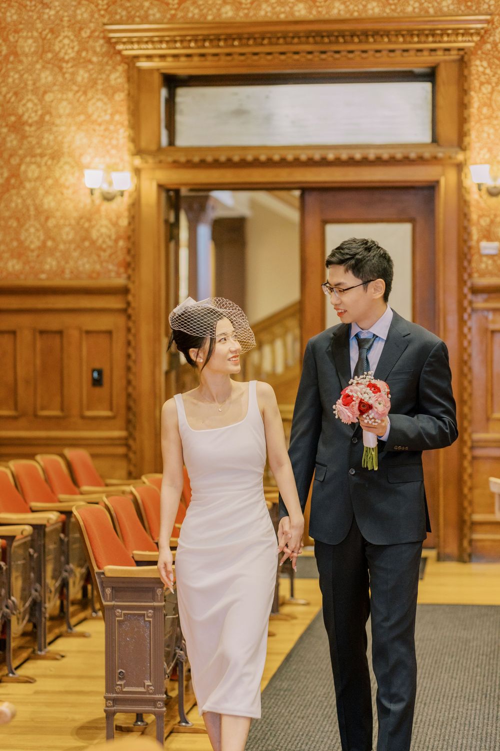 Newlyweds walking through a grand hallway with vintage wooden walls and coral-colored chairs.