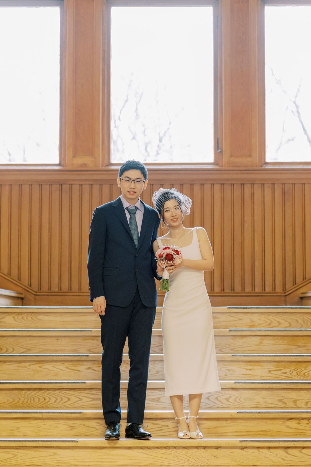 A couple in wedding attire stands on wooden stairs in formal indoor setting.