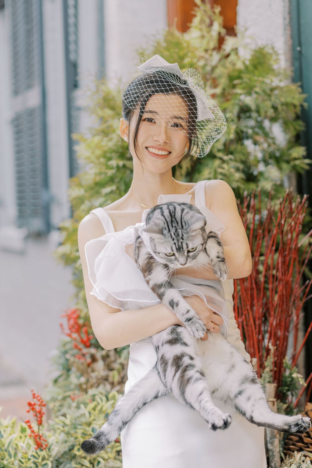 A bride wearing a birdcage veil holds a large grey and white cat in front of a garden setting.
