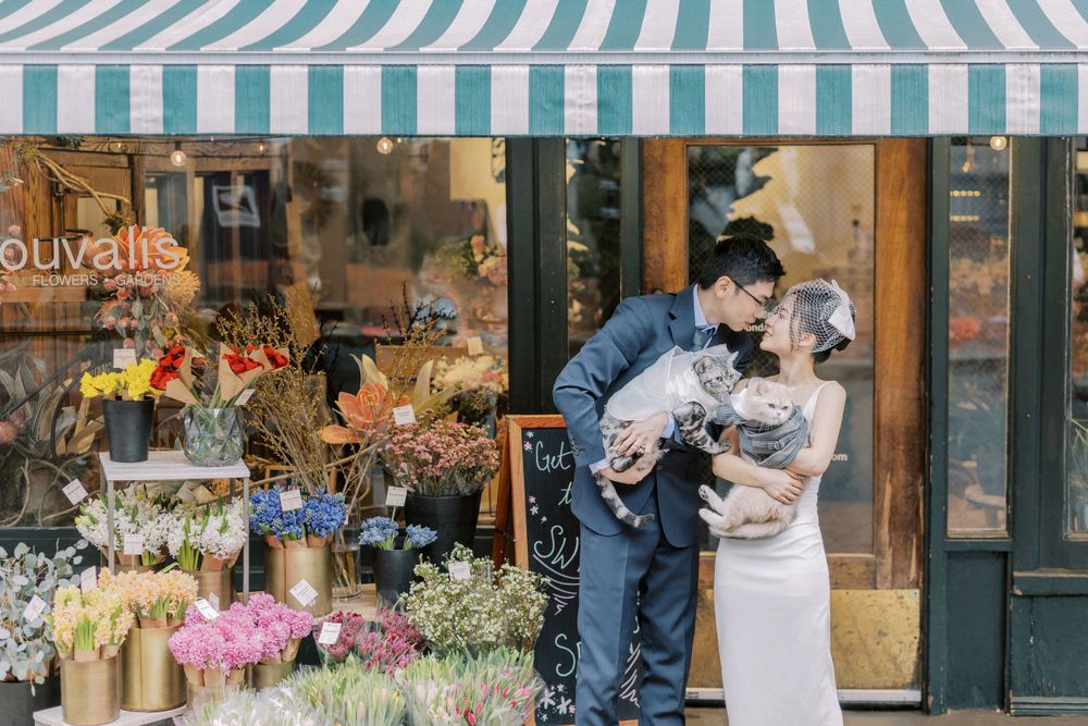 A couple with their cat pose in front of a flower shop with a striped awning.