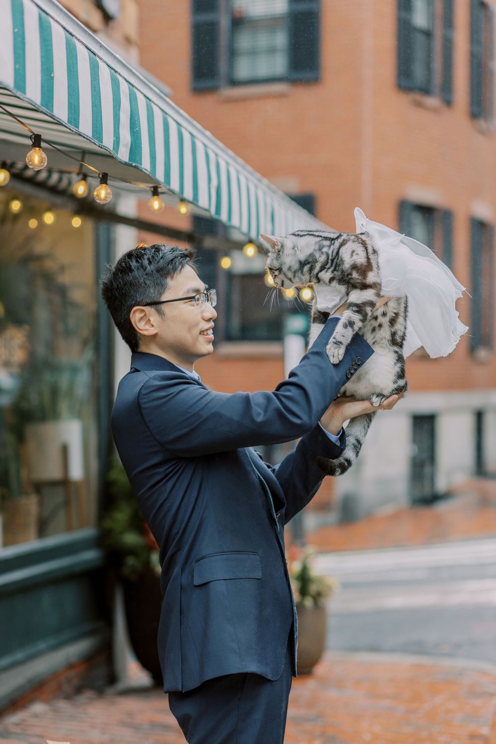A groom in navy suit lifts a cat wearing a white veil against a brick building backdrop.