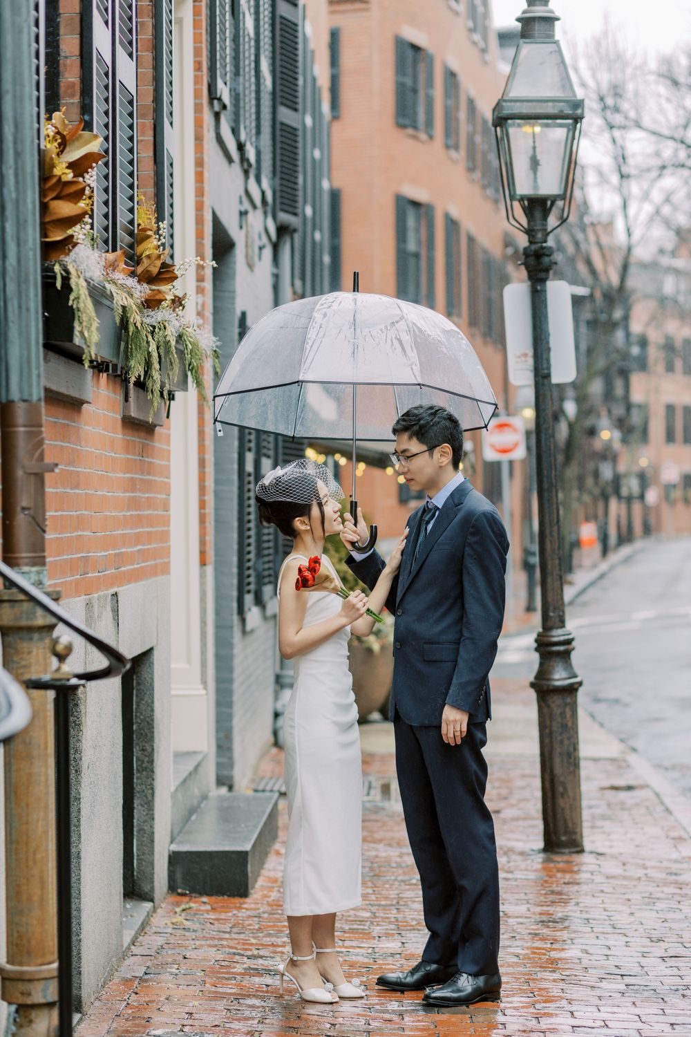 A couple shares an umbrella on a rainy cobblestone street near vintage lampposts.