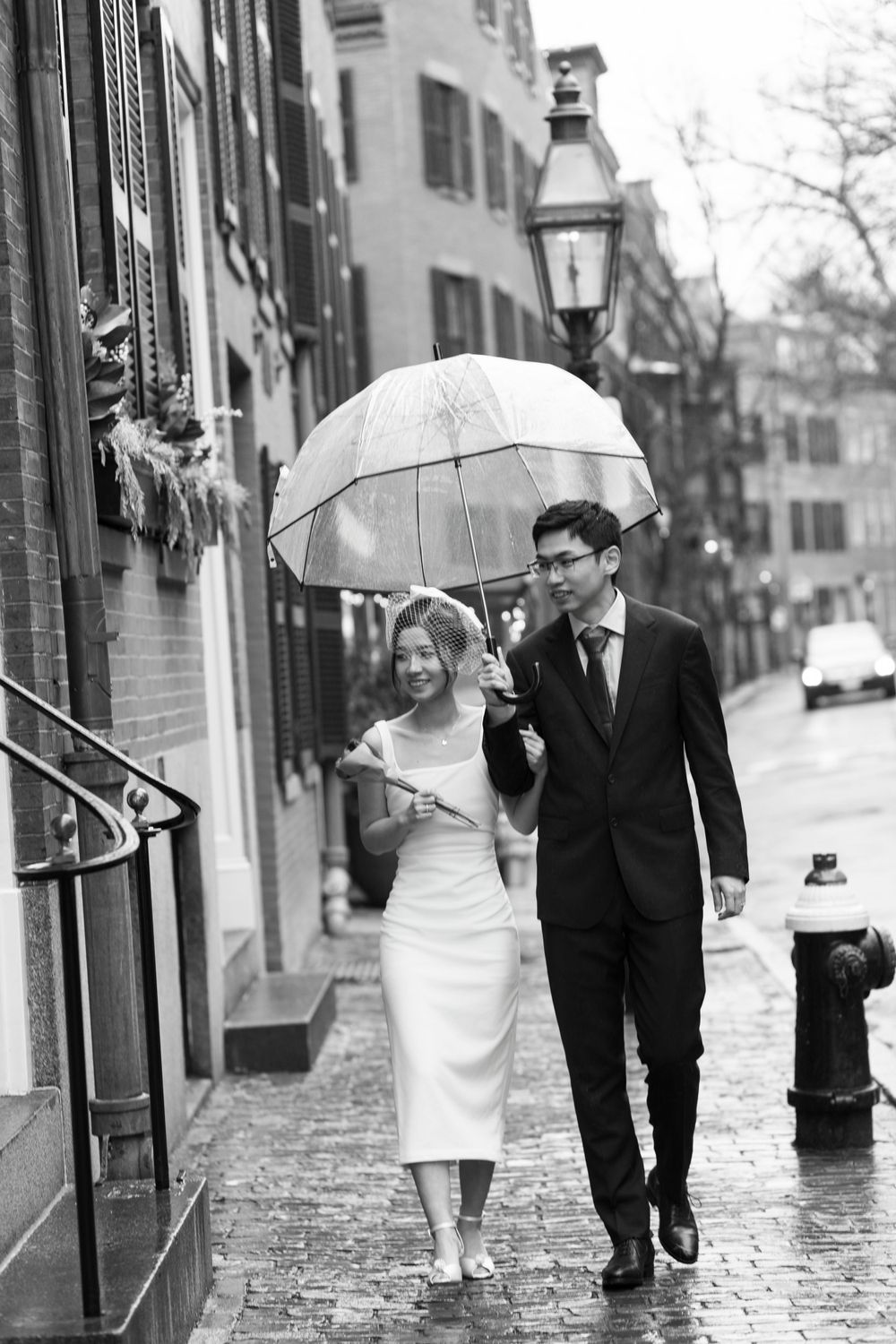 A couple shares an umbrella while walking on a rainy cobblestone street in black and white.