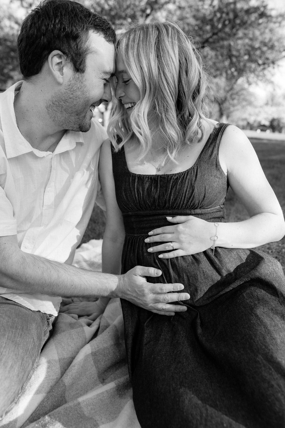 Black and white maternity photo of a couple sitting outdoors, tenderly touching the pregnant woman's belly.