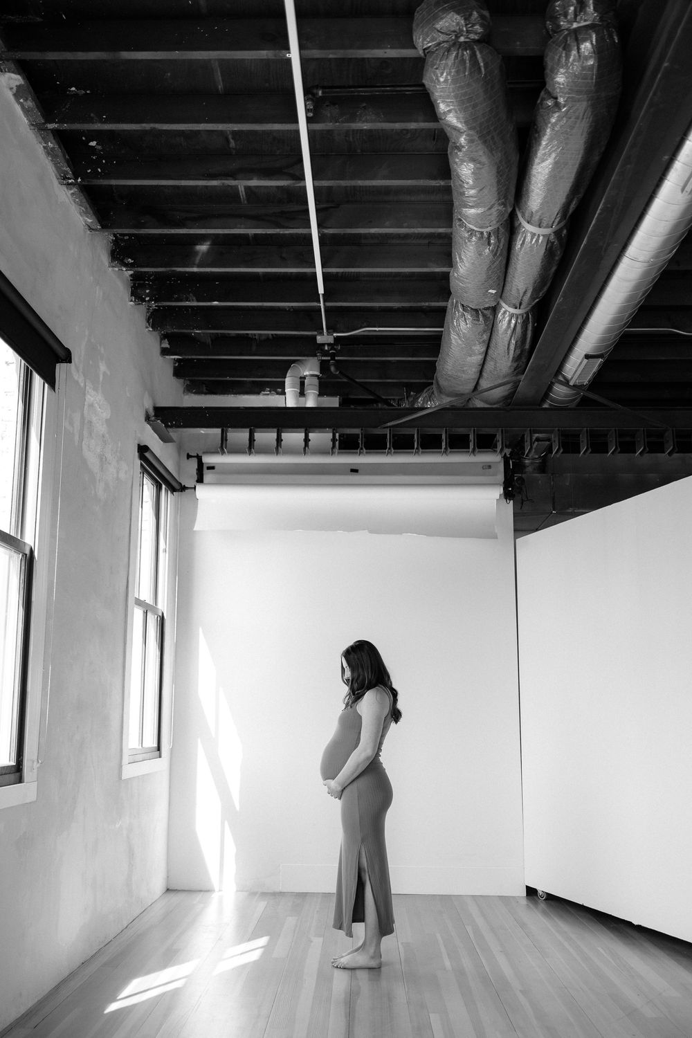 Black and white portrait of an expectant mother standing in an empty room with exposed industrial ceiling and sunlit windows.