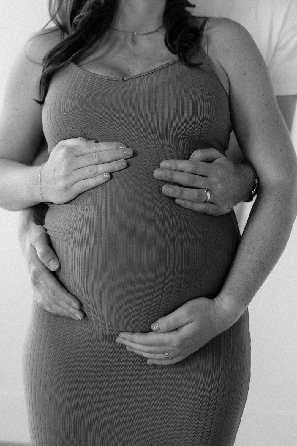 Black and white photo of hands gently cradling a pregnant belly in a ribbed maternity dress.