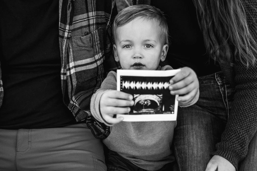 Small child holds ultrasound photo while sitting between adults. Family photo session.