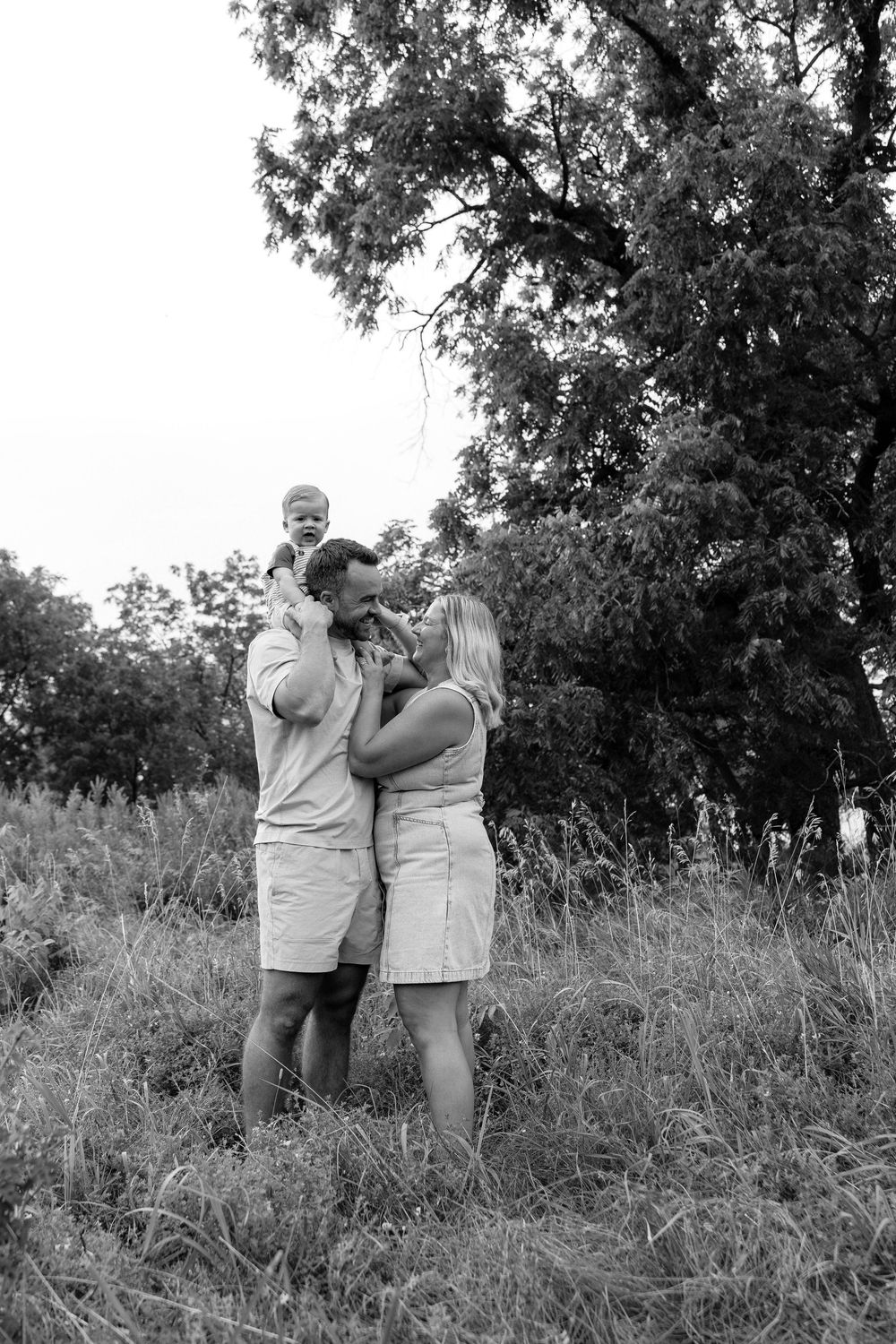 Two people in light clothing share a tender moment while standing in a grassy field surrounded by trees.