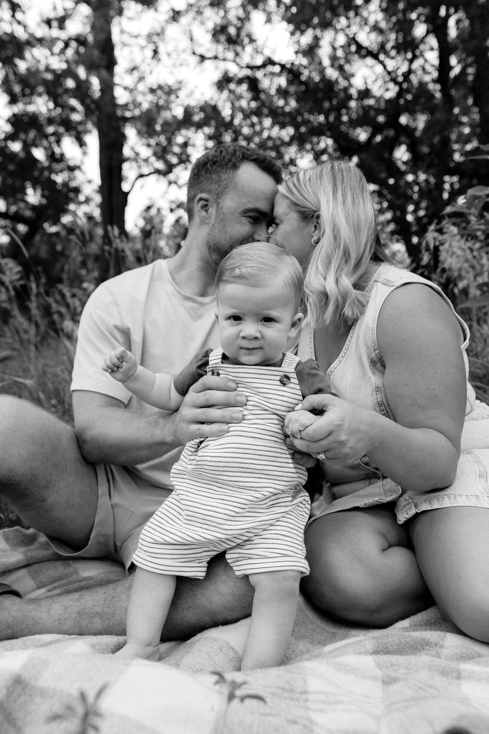 A young family shares an intimate moment together on a picnic blanket in a wooded setting captured in black and white.