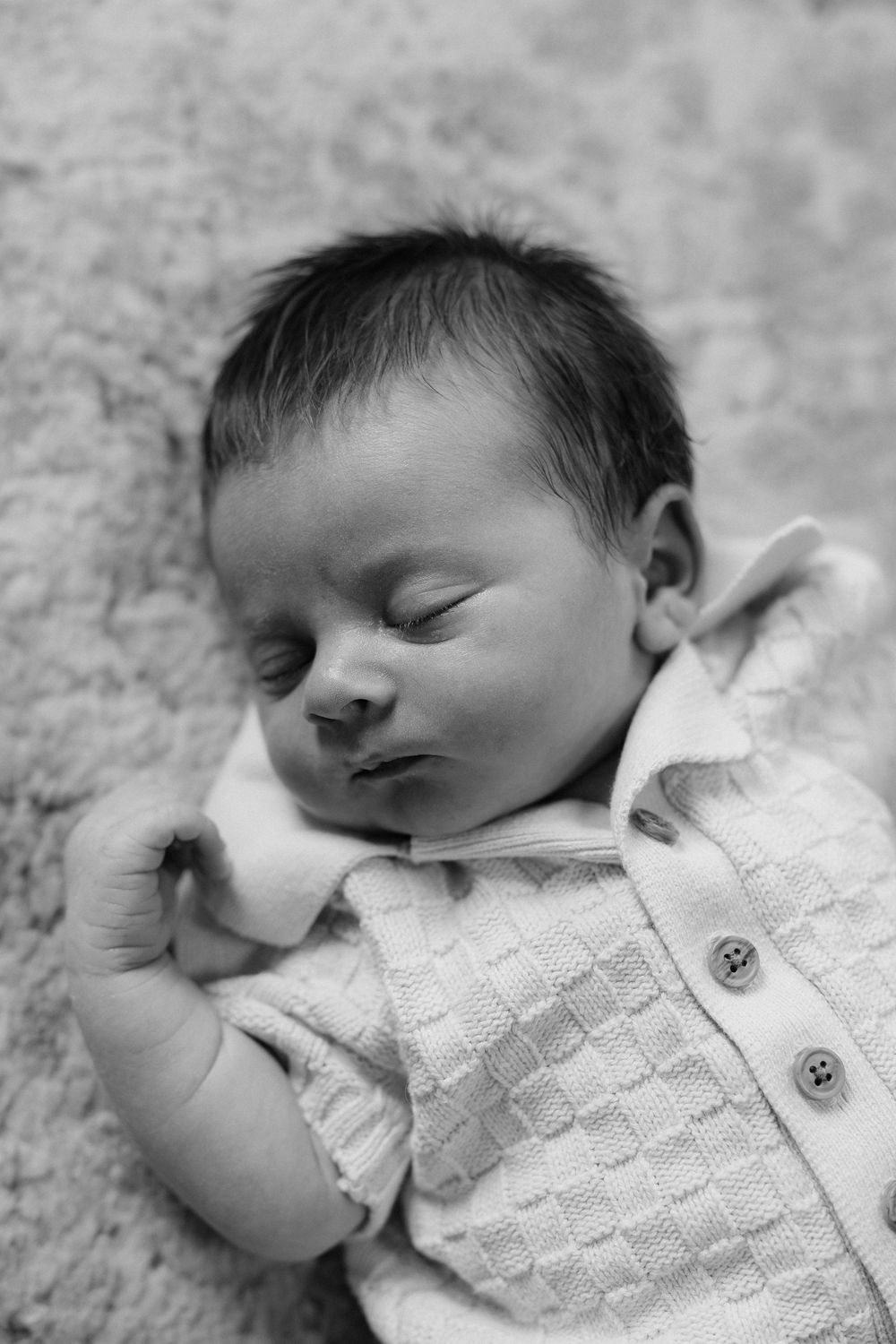 A sleeping newborn in a white knit cardigan rests peacefully against a textured blanket in black and white.