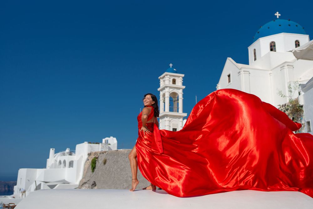 Dramatic red dress flows against white architecture and blue dome of classic Santorini church.