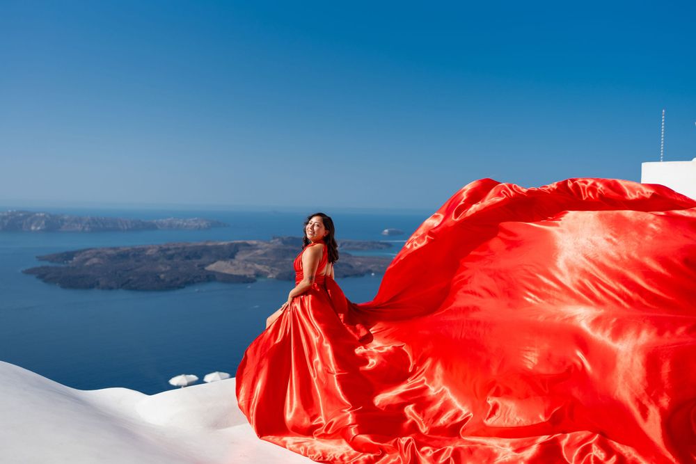 Dramatic red gown billowing in the wind against Santorini's iconic white buildings and sea views.