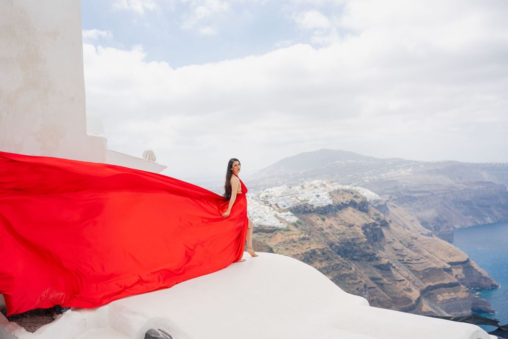 Flowing red dress creates striking contrast against white buildings and Aegean Sea views.